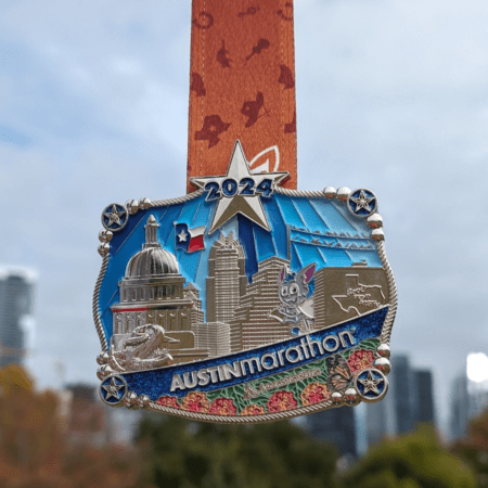 A 2024 Austin Marathon medal featuring a detailed cityscape of Austin landmarks, including the Texas State Capitol, with colorful enamel accents and a star at the top. The belt buckle medal hangs from an orange ribbon adorned with silhouettes of runners against a cloudy sky backdrop. Austin Marathon Half Marathon & 5K