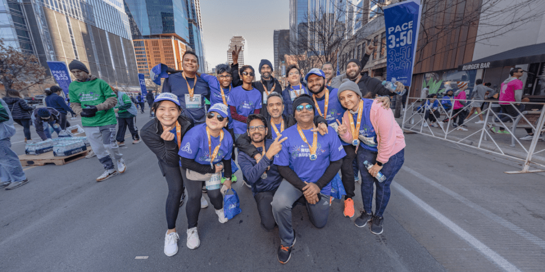 A group of happy marathon runners, all wearing blue T-shirts, pose together on a city street at the Finish Line Festival. Some are holding medals and making peace signs. Skyscrapers and other participants can be seen in the background. A PACE 3:50 sign is visible in the distance. Austin Marathon Half Marathon & 5K