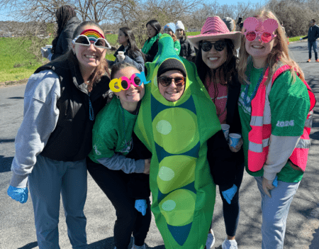 A group of five people smiles for the camera outdoors. One person is wearing a pea pod costume, and the others are dressed in various colorful outfits with fun accessories like large sunglasses and hats. Behind them, aid stations are set up among the trees where other people can be seen enjoying the day. Austin Marathon Half Marathon & 5K