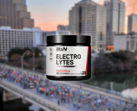 A black jar labeled "Electrolytes" is prominently displayed in the foreground. The background shows a cityscape with tall buildings and a blurry crowd, possibly at an aid station during a marathon or event on a bridge. The label indicates it provides hydration with 50 servings. Austin Marathon Half Marathon & 5K