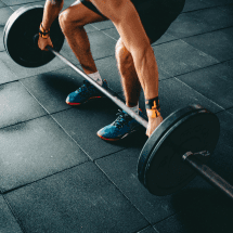 A person is bent over, gripping a barbell with both hands, preparing to lift heavy weights from the floor. The individual is wearing athletic wear, including shorts, a wristband, and blue and red runners, standing on a black rubber gym floor. Austin Marathon Half Marathon & 5K