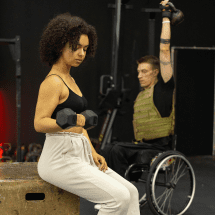 A woman sitting on a wooden box lifts light weights while a man in a wheelchair, wearing a weighted vest, raises a kettlebell behind her. The gym setting includes exercise equipment and a dark background. Austin Marathon Half Marathon & 5K