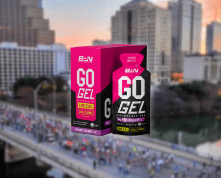 A product image of BPN Go Gel in mixed berry flavor. The box and gel packet are prominently displayed, featuring text highlighting "100 CAL" and "24G CARBS." The background shows an urban marathon scene with runners crossing a bridge while aid stations dot the course and city buildings tower above. Austin Marathon Half Marathon & 5K