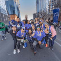 A community of smiling runners wearing blue shirts and medals pose for a photo on a city street after a race. Skyscrapers and other runners can be seen in the background. A "Pace 3:50" sign is visible on the right side. Austin Marathon Half Marathon & 5K