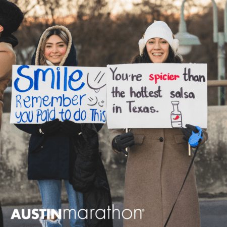 Two people stand outdoors, bundled up in warm clothing, holding humorous signs. One sign says, "Smile. Remember you paid to do this," while the other reads, "You're spicier than the hottest salsa in Texas." Both are smiling. Text reads "Austin Marathon" at the bottom—a celebration of marathon running myths and camaraderie. Austin Marathon Half Marathon & 5K