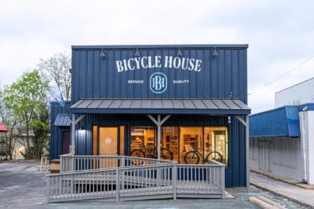 A blue-painted storefront with a sign reading "Bicycle House" and "Service Quality." The shop features large windows showing bicycles inside, perfect for both casual cycling and marathon training. A ramp leads up to the entrance, and the exterior integrates metallic and wooden elements. Trees and neighboring buildings are visible. Austin Marathon Half Marathon & 5K