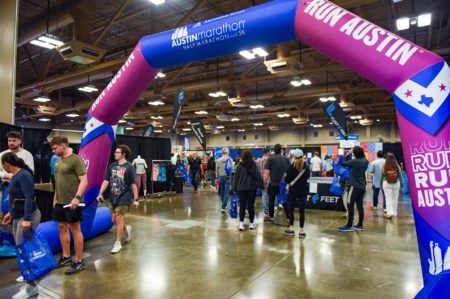 People visit a convention center for the Austin Marathon, with booths and merchandise displays around. An archway labeled "RUN AUSTIN" stands in the foreground, while attendees browse and interact in the background. Businesses are encouraged to become a sponsor to gain visibility among participants. Austin Marathon Half Marathon & 5K