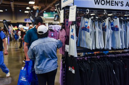 Two people are shopping at a merchandise stall in a convention center. They are browsing a rack of shirts with "Austin" written on them. The background shows other stalls and people walking around, some passing by a sign encouraging attendees to become a sponsor. The hanging sign reads "Women's Apparel. Austin Marathon Half Marathon & 5K