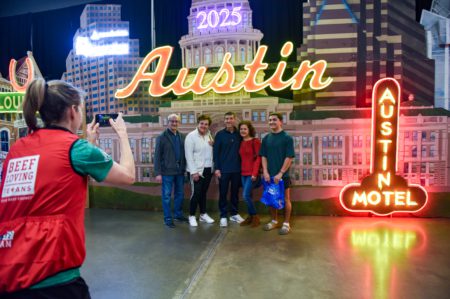 A person in a red vest takes a photo of five people smiling and posing together in front of a brightly lit "Austin" sign with cityscape elements like the Austin Motel sign and other buildings. The year "2023" is displayed at the top, encouraging visitors to become a sponsor for further events. Austin Marathon Half Marathon & 5K