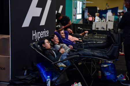 Four people are sitting in reclining chairs with their legs elevated in Hyperice compression therapy devices. They appear relaxed and are smiling. The background features a Hyperice banner and some booths, with people and products visible in the distance—an inviting scene for those looking to become a sponsor. Austin Marathon Half Marathon & 5K