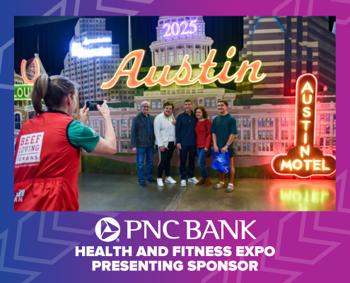 A group of people poses for a photo in front of an illuminated "Austin" sign at an indoor event featuring the Austin Marathon. The backdrop showcases the city's skyline, while a PNC Bank banner proudly reads, "Health and Fitness Expo Presenting Sponsor. Austin Marathon Half Marathon & 5K