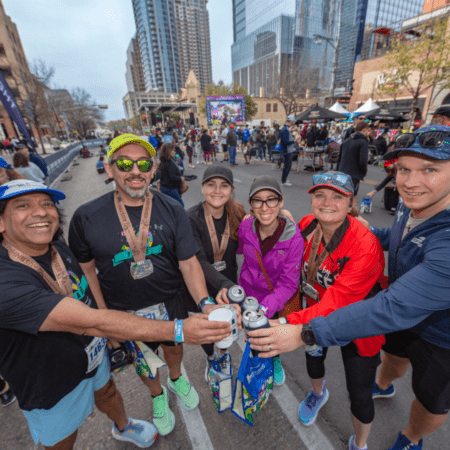 A group of six people in athletic gear celebrate with canned drinks on a city street during the 2025 Austin Marathon. They wear medals around their necks, surrounded by tall buildings and other jubilant race day participants. The mood is joyful and festive. Austin Marathon Half Marathon & 5K