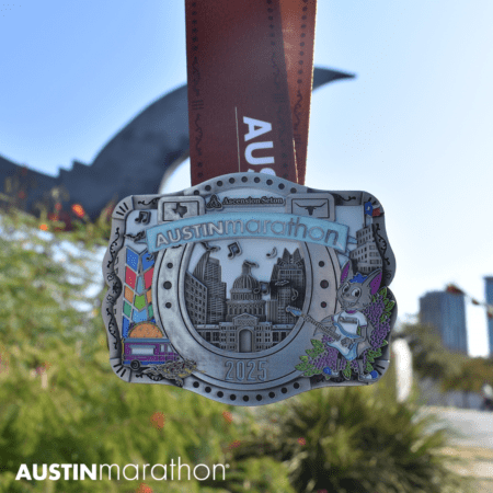 Close-up of the 2025 Austin Marathon finisher medal, showcasing a detailed cityscape with iconic buildings and landmarks. The brown ribbon reads “AUSTINmarathon.” In the slightly blurred background, lush greenery and a city skyline shimmer, enhancing the allure of this exceptional race bling. Austin Marathon Half Marathon & 5K