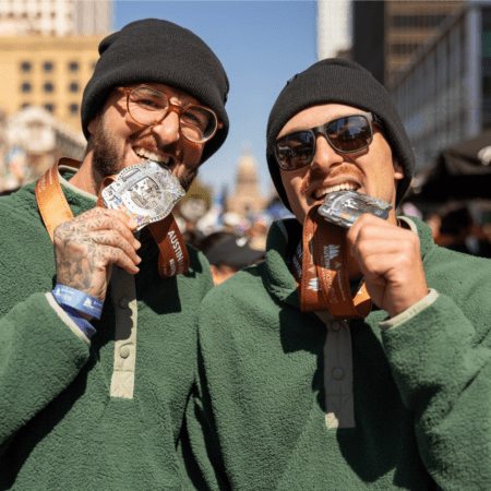 Two smiling runners in green fleeces and black beanies proudly bite medals around their necks, celebrating at a marathon in Austin with buildings and a lively crowd in the background. Austin Marathon Half Marathon & 5K