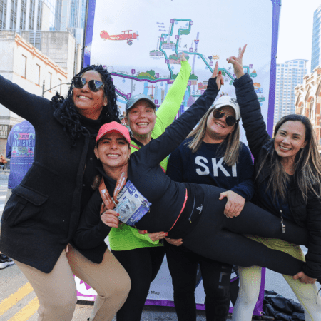 Five women, joyful runners, strike a playful pose in front of a colorful marathon map at the Marathon in Austin. One is lifted horizontally by her friends—all wear casual athletic clothing, hats, and sunglasses, looking happy and celebratory. Austin Marathon Half Marathon & 5K