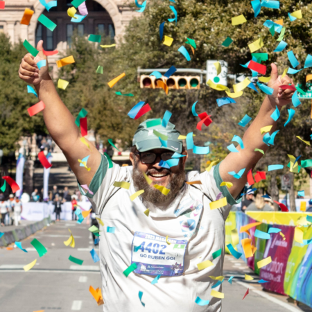 A smiling runner with a beard and glasses raises his arms in celebration as colorful confetti falls around him at the Marathon in Austin finish line. He wears a cap, sports shirt, and bib number 4402, embodying the spirit of all dedicated runners. Austin Marathon Half Marathon & 5K