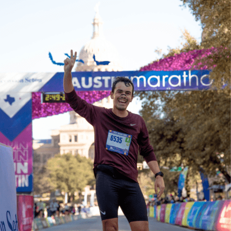 A smiling runner raises a peace sign as he crosses the finish line at the best marathon in Austin, with race bib 6535. The event banner and a dome-shaped building are visible in the background. Austin Marathon Half Marathon & 5K