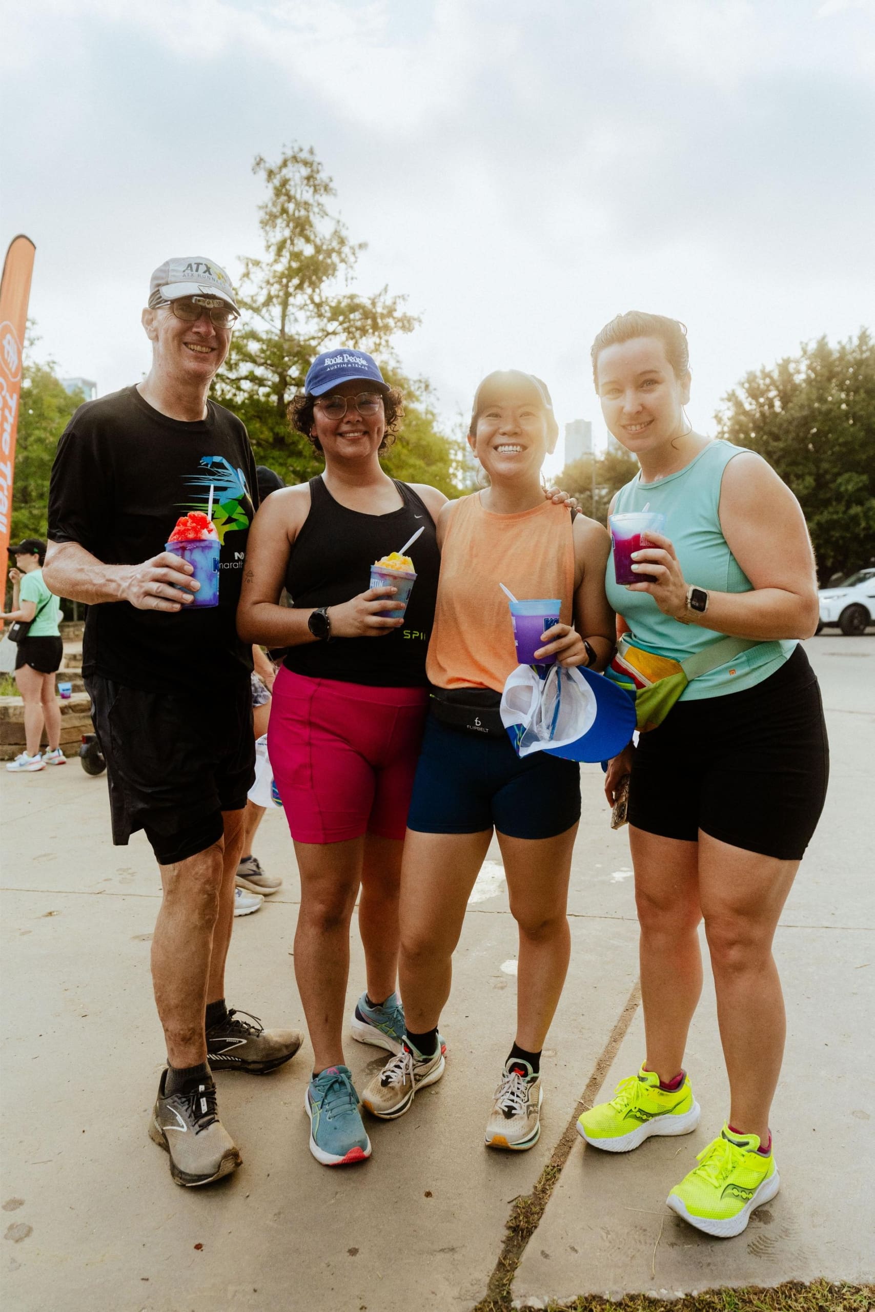 Four people in athletic wear stand outdoors, smiling and holding colorful drinks—Snow Cones on Trail—suggesting they just finished a run or race. Trees and fellow participants can be seen in the background. Austin Marathon Half Marathon & 5K
