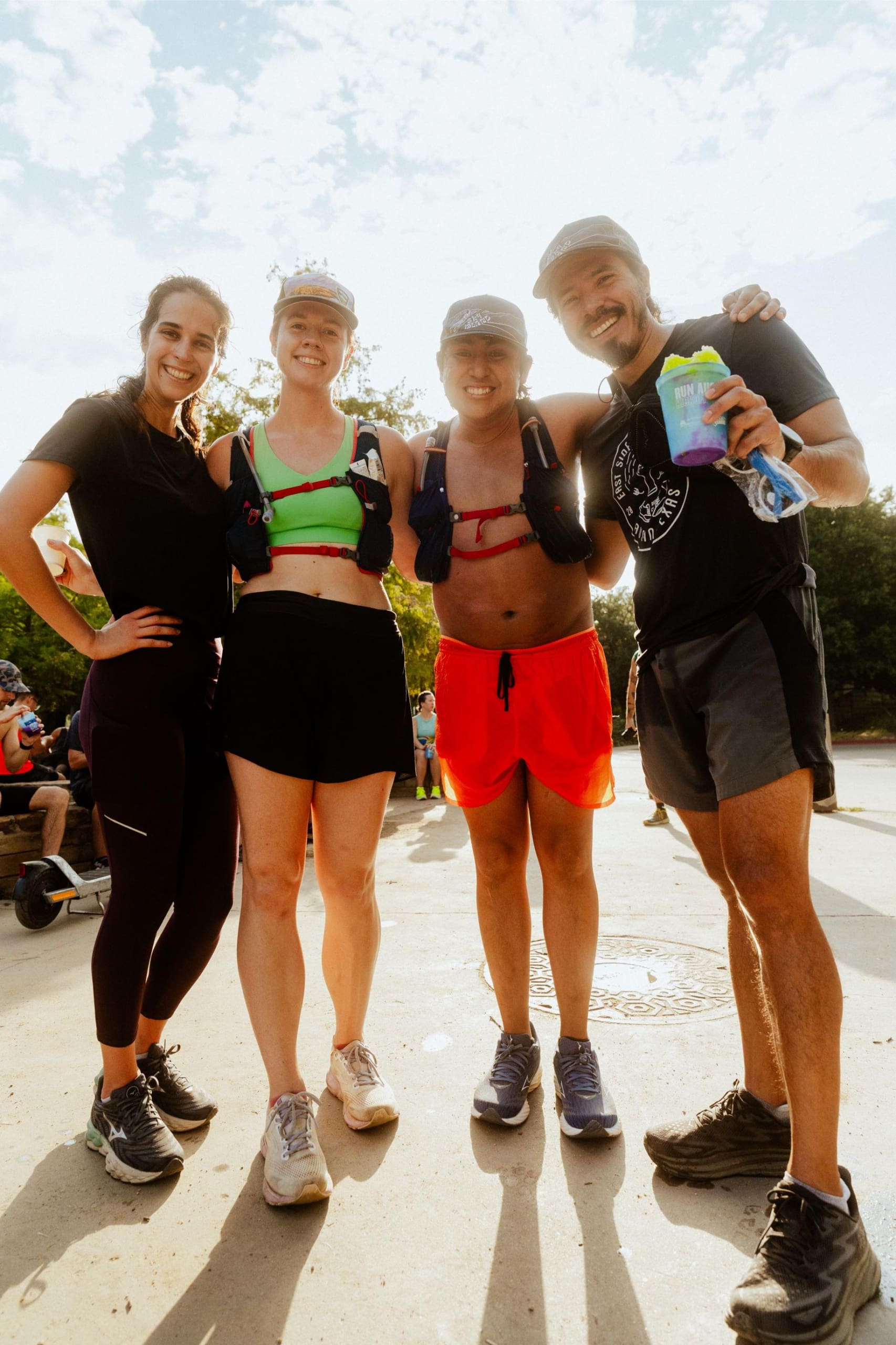 Four smiling people in athletic wear stand close together outdoors on a sunny day, appearing happy after a run or workout—perhaps celebrating with snow cones on trail. Trees and other people are visible in the background. Austin Marathon Half Marathon & 5K