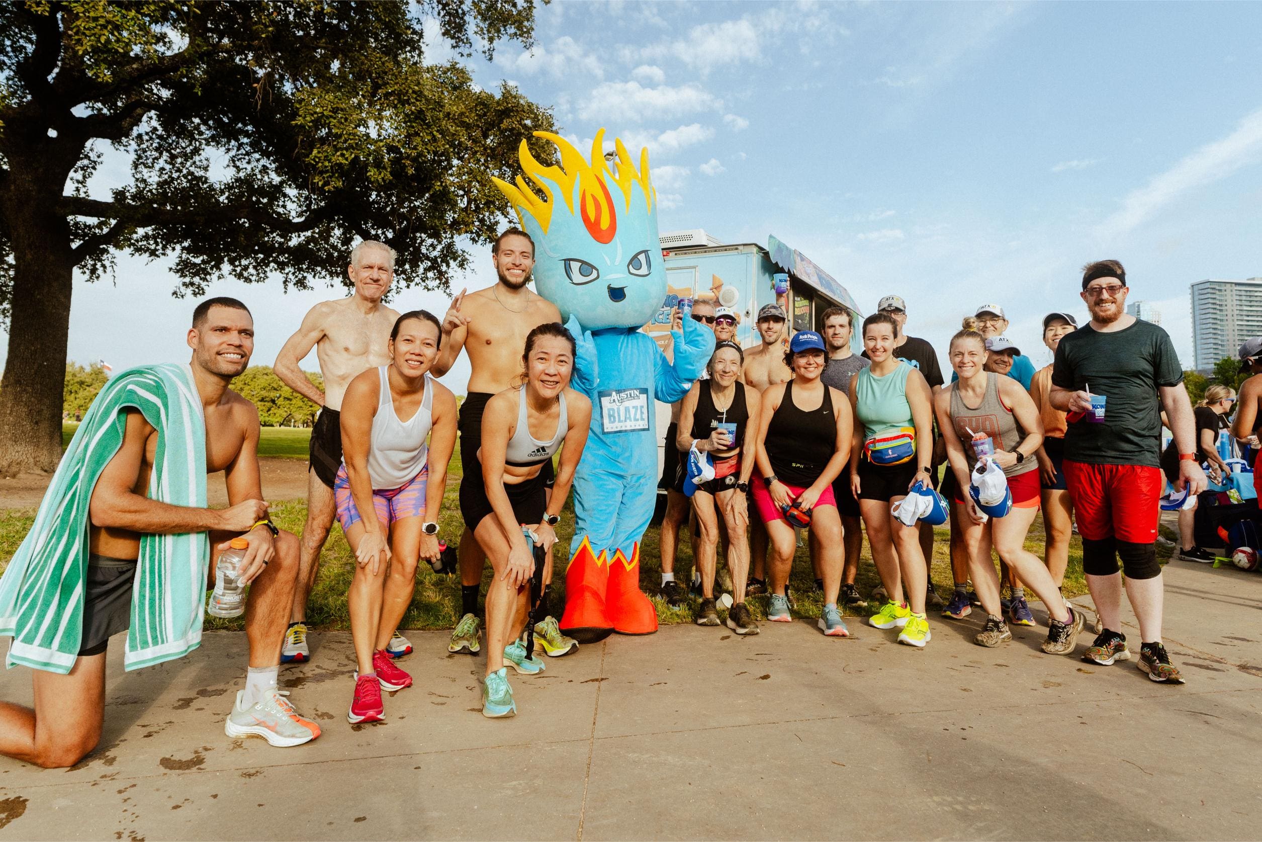 A group of smiling runners poses outdoors with a person in a blue flame mascot costume. Clutching water bottles and enjoying Snow Cones on Trail, they stand in athletic gear with trees and buildings in the background on a sunny day. Austin Marathon Half Marathon & 5K