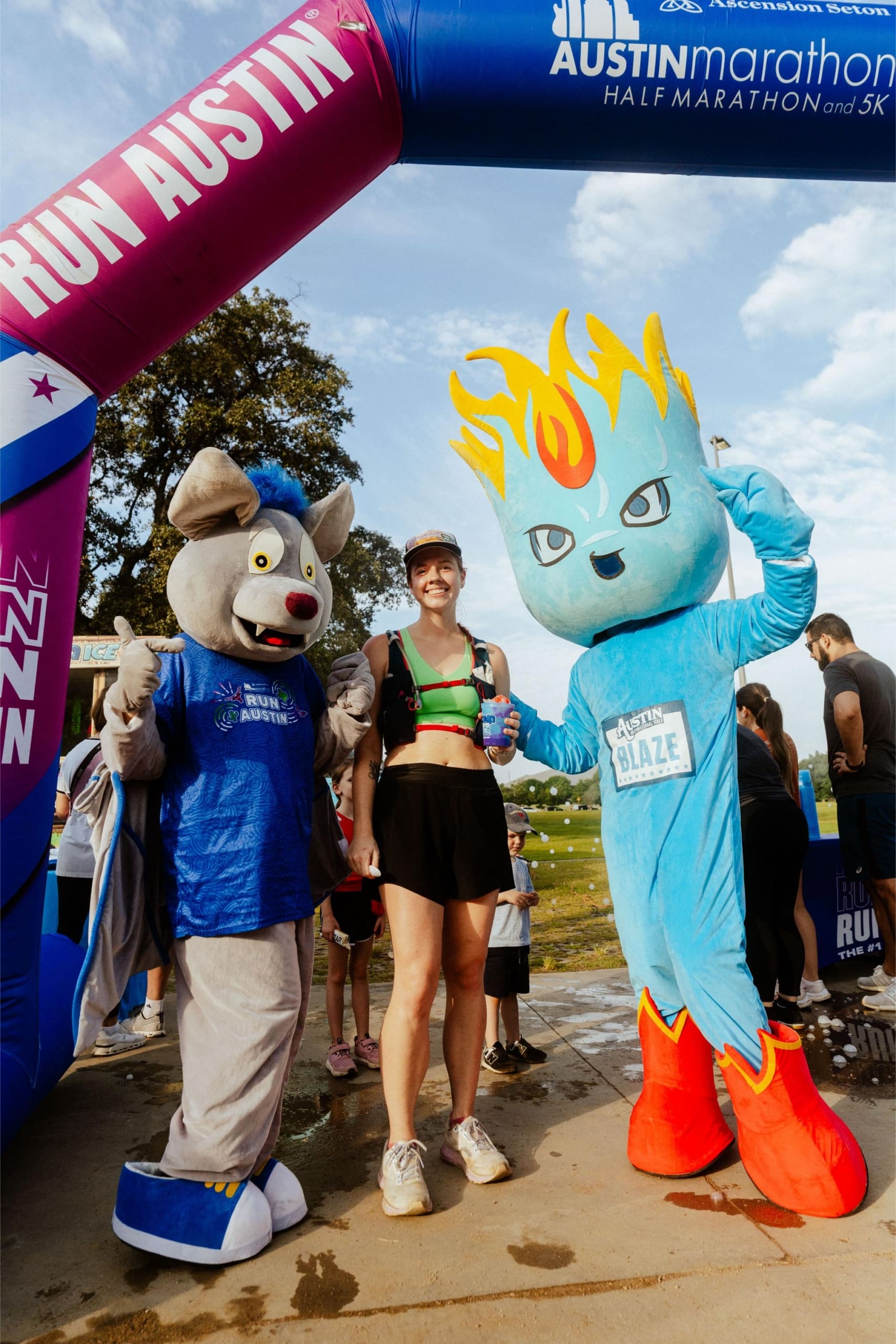A smiling runner stands between two colorful mascots at the finish line of the Austin Marathon, under an arch that reads "RUN AUSTIN," with Snow Cones on Trail nearby. Other people and trees are visible in the background. Austin Marathon Half Marathon & 5K