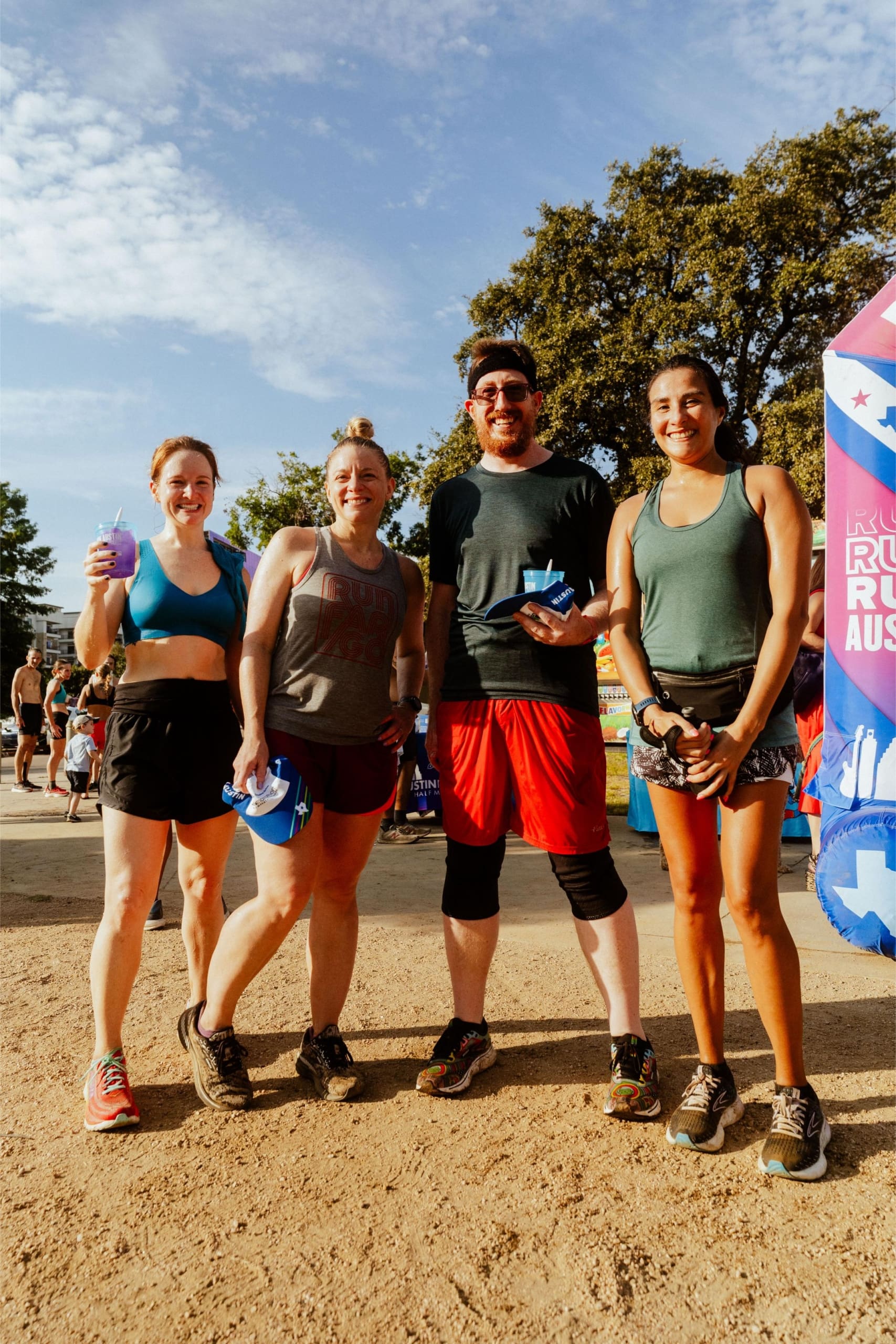 Four runners stand smiling together outdoors at a race event, holding water bottles. They wear athletic clothes and sneakers, with trees, a blue sky, and Snow Cones on Trail banners visible among other participants in the background. Austin Marathon Half Marathon & 5K