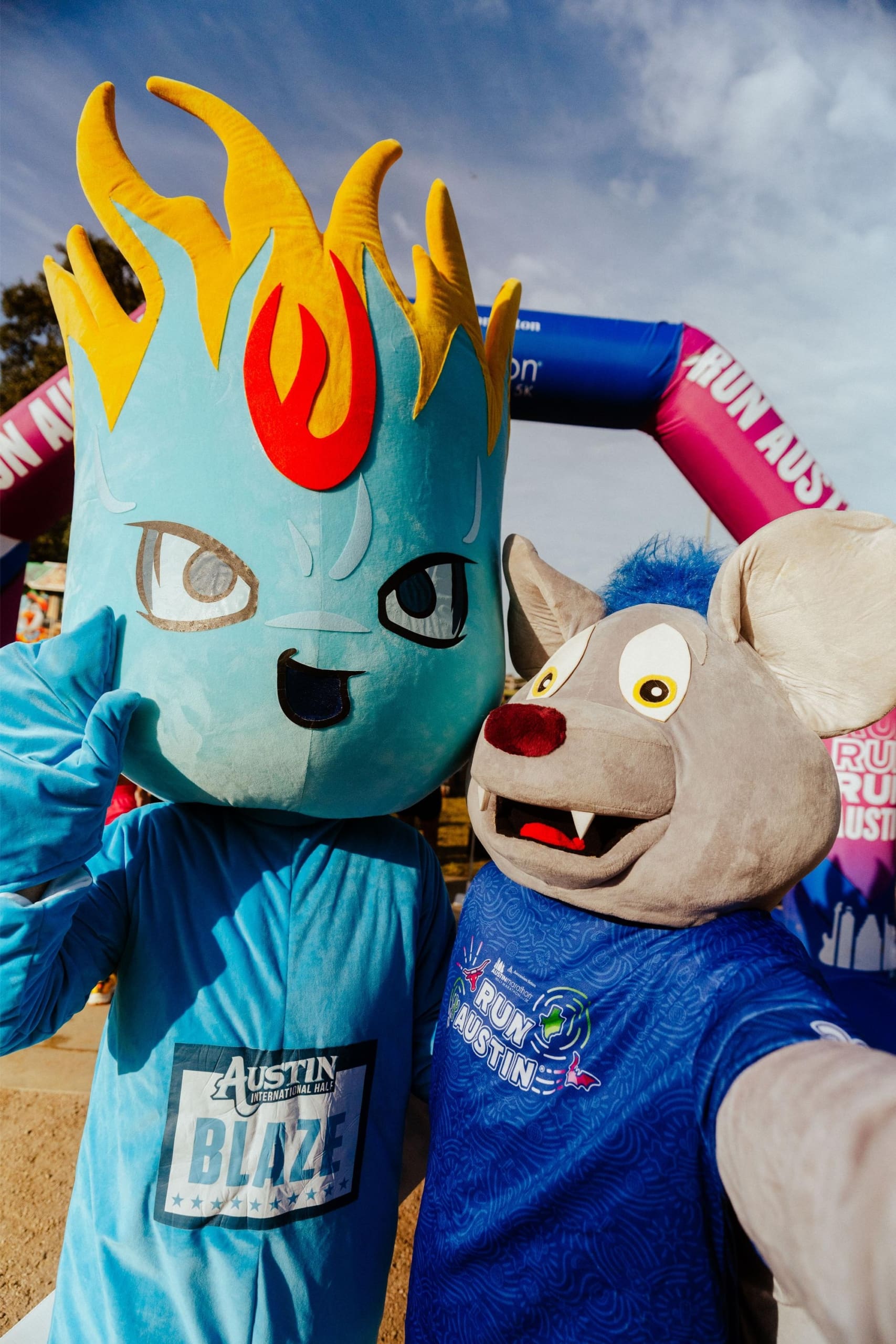 Two mascots in costume pose for a selfie at an outdoor event, with vibrant arches behind them. The blue flame-haired mascot and the gray mouse in a blue shirt celebrate the fun of Snow Cones on Trail. Austin Marathon Half Marathon & 5K