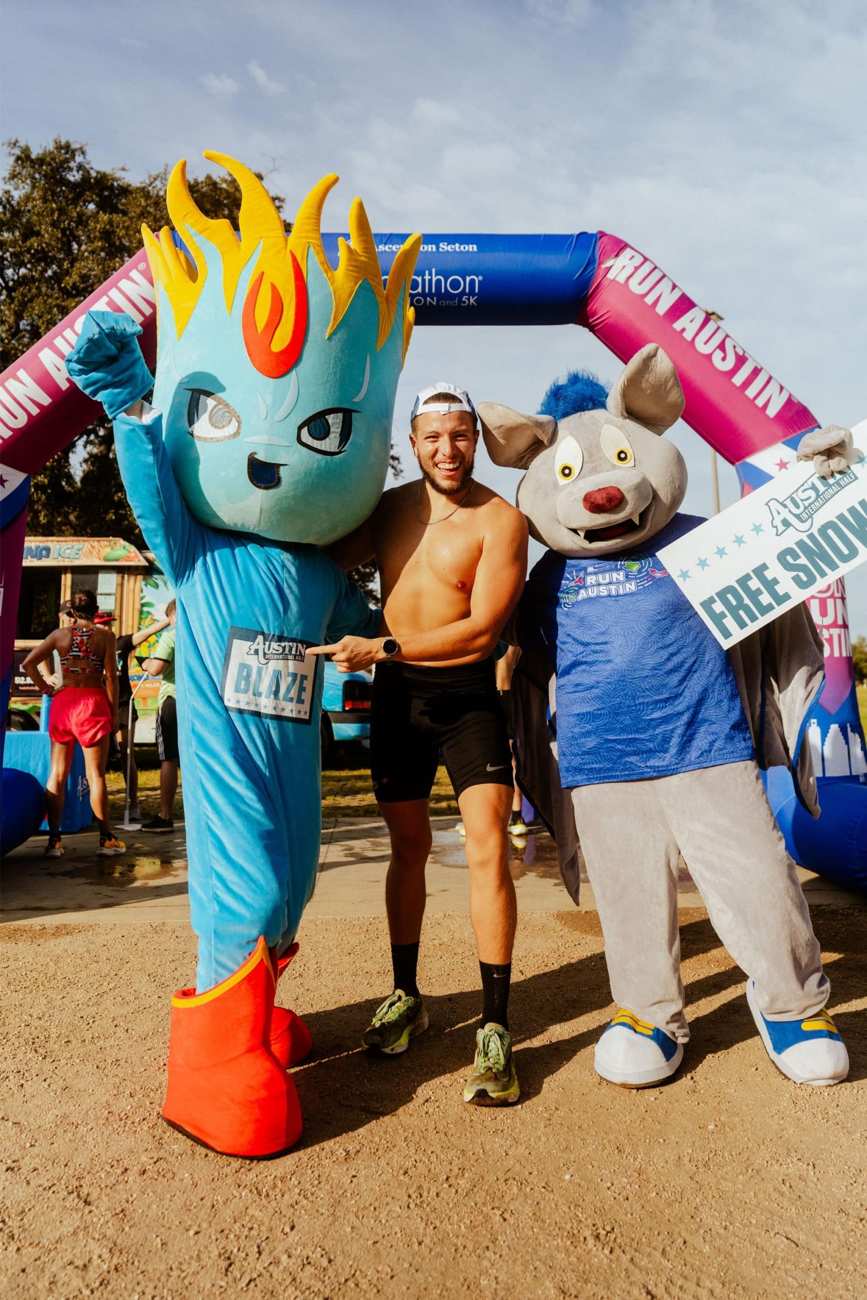 A smiling runner poses between two mascots in costume at a race finish line, celebrating with Snow Cones on Trail. One mascot is blue with a flame head, the other is a gray bat holding a "FREE SQUAD" sign. An inflatable arch and trees are in the background. Austin Marathon Half Marathon & 5K