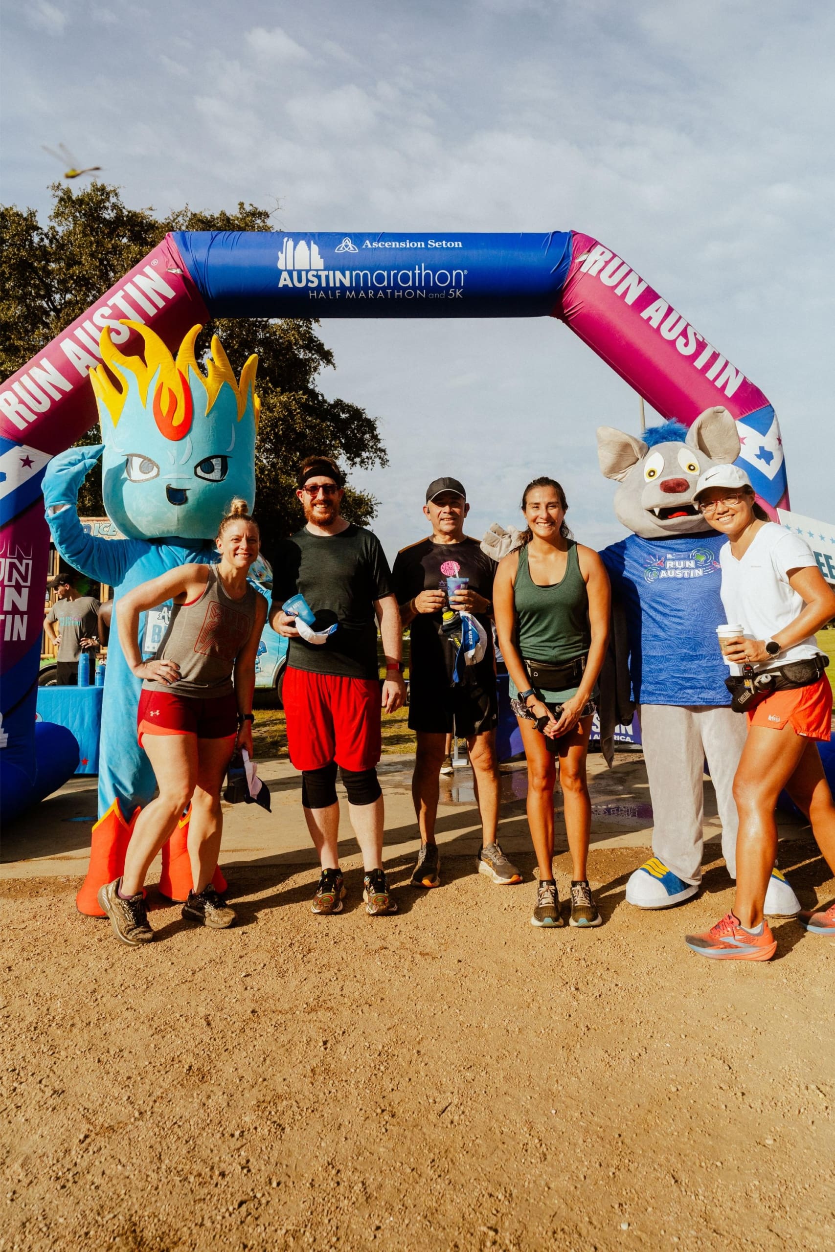 A group of marathon runners pose smiling under an Austin Marathon archway, joined by two mascots—one a blue flame character, the other a gray animal in a blue shirt—with Snow Cones on Trail stands nearby. The sun is shining and trees fill the background. Austin Marathon Half Marathon & 5K