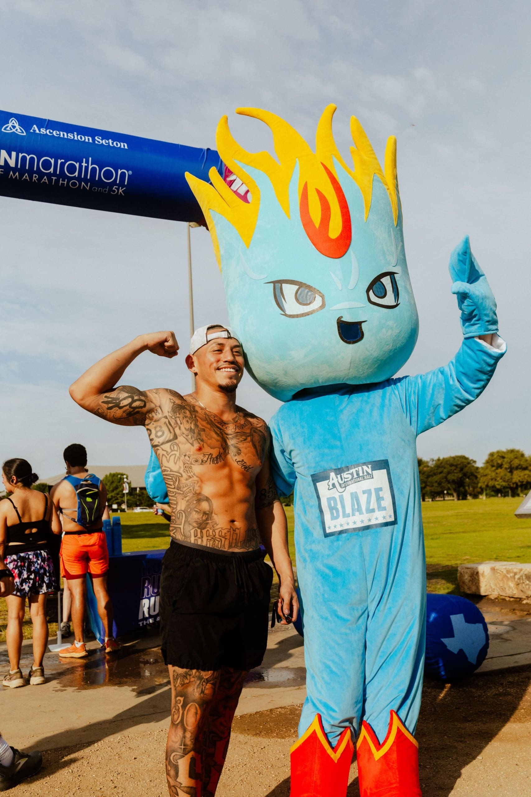 A shirtless, tattooed man flexes his arm and smiles beside the Austin Blaze mascot—blue-bodied with fiery yellow hair—at a marathon event finish line, where participants celebrate and enjoy Snow Cones on Trail in the background. Austin Marathon Half Marathon & 5K