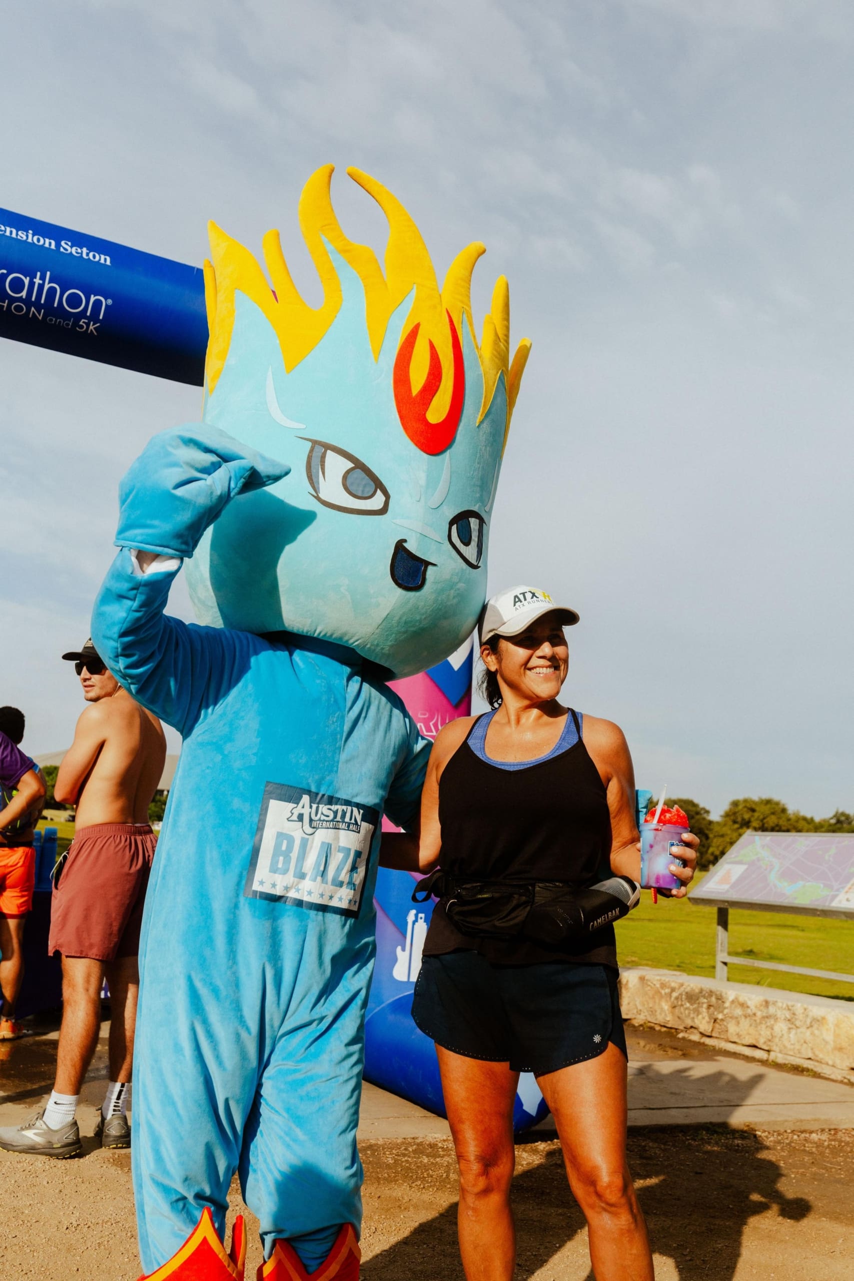 A person in a flame-headed blue mascot costume poses with a smiling woman in athletic gear near the marathon finish line, where runners enjoyed Snow Cones on Trail at the outdoor event. Austin Marathon Half Marathon & 5K