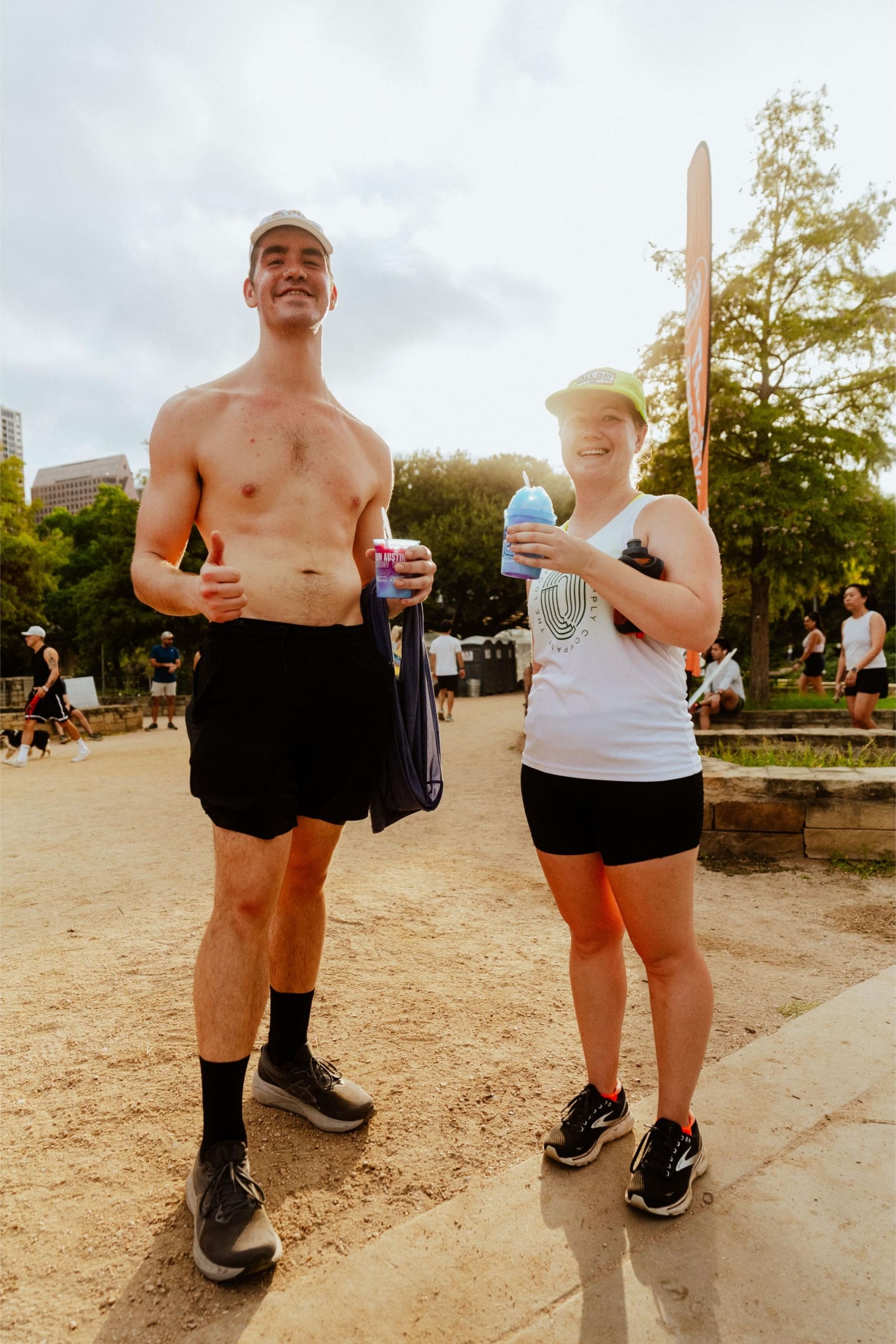 A man and woman in athletic wear stand outdoors on a dirt path, smiling and holding snow cones on trail. The man is shirtless, and both wear hats. Other people and trees are visible in the background on a sunny day. Austin Marathon Half Marathon & 5K