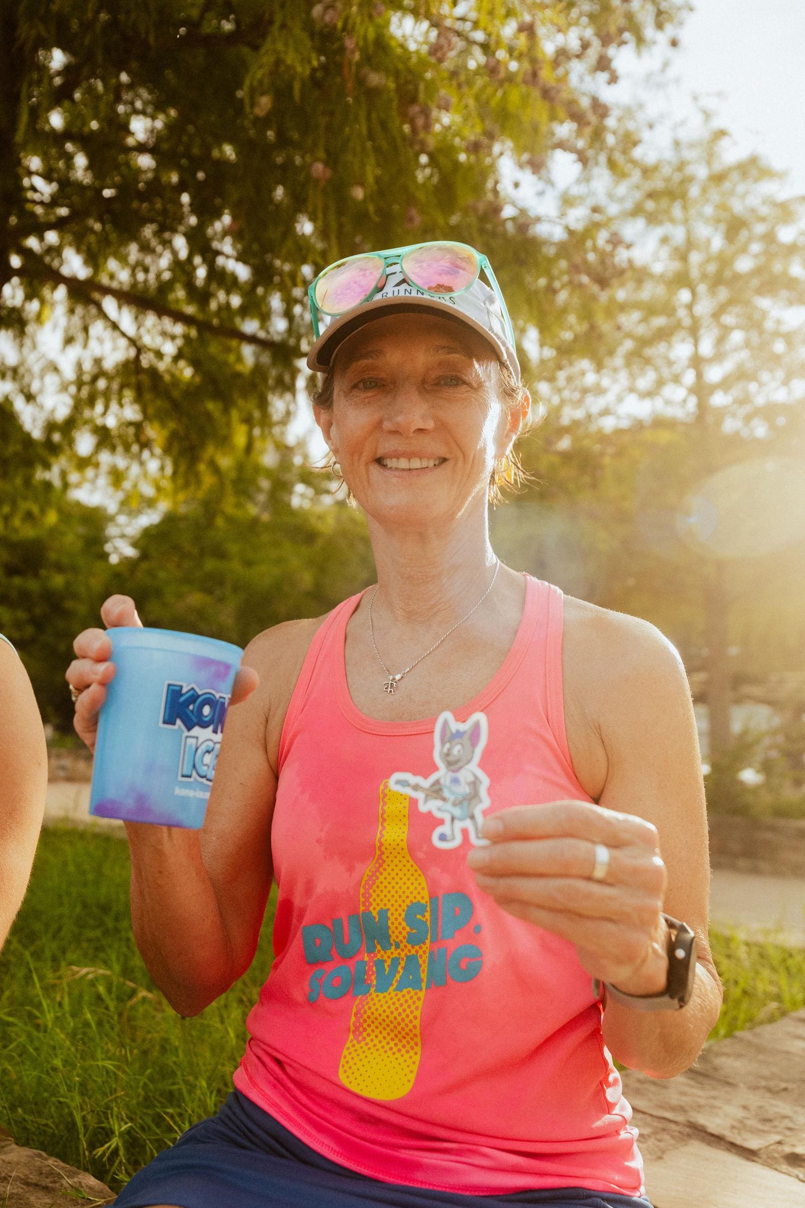 A smiling woman in a pink tank top and cap holds a blue cup and a small cartoon cat sticker, enjoying Snow Cones on Trail outdoors on a sunny day. She wears sunglasses on her head and sits on grass with trees behind her. Austin Marathon Half Marathon & 5K