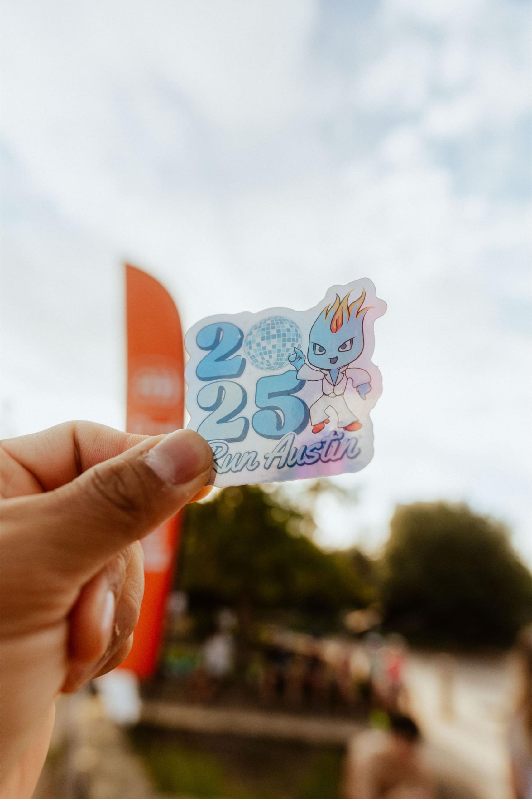 A hand holds a sticker that reads “2025 Run Austin” with a cartoon character and disco ball graphic, against a blurred outdoor background with trees, a red vertical banner, and hints of Snow Cones on Trail festivities. Austin Marathon Half Marathon & 5K