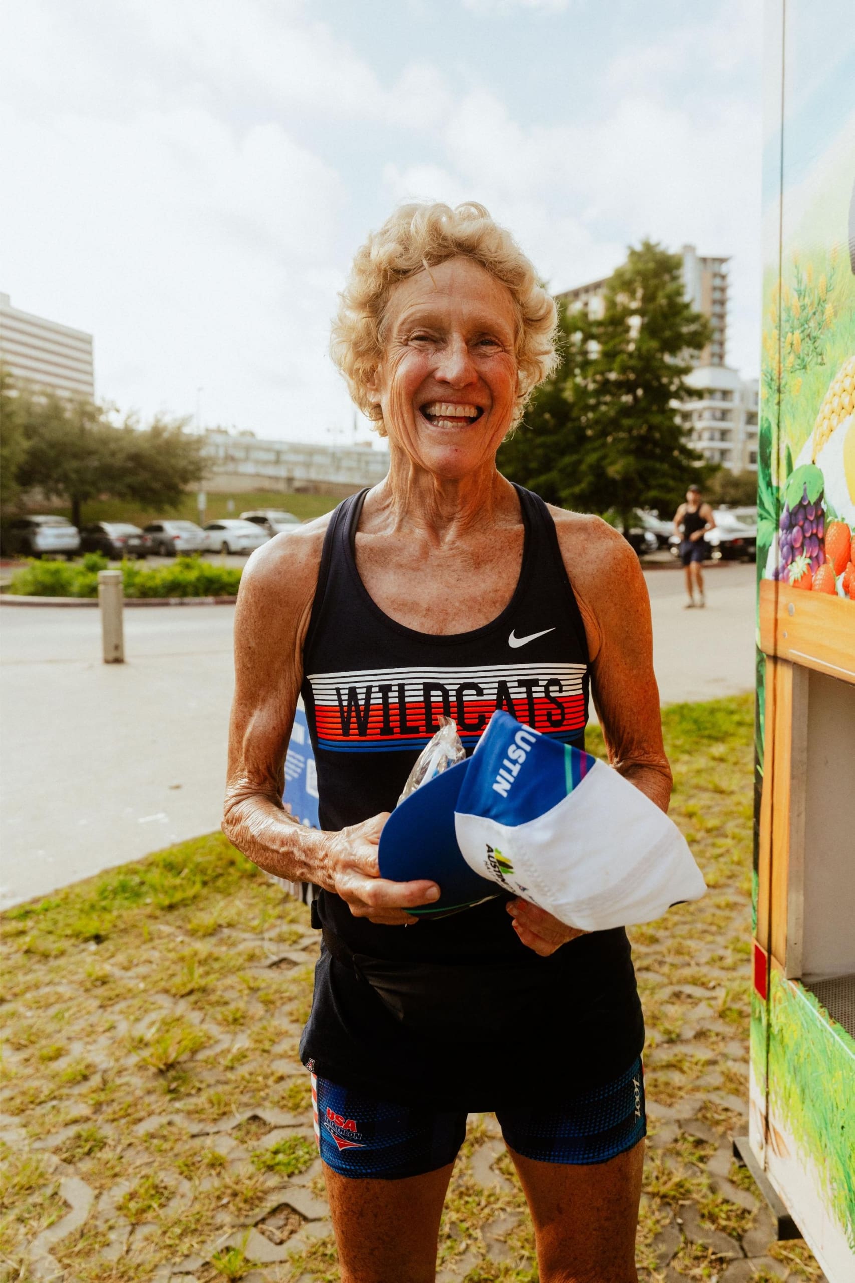 An older woman with curly blonde hair, wearing a "WILDCATS" running tank, smiles while holding a blue and white cap outdoors on a sunny day, enjoying Snow Cones on Trail with buildings and greenery in the background. Austin Marathon Half Marathon & 5K