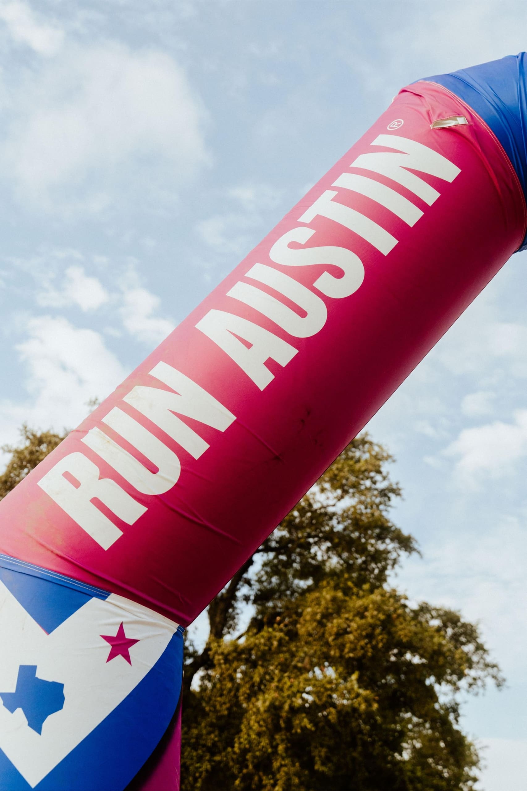 A large pink and blue inflatable arch with the words "RUN AUSTIN" in bold white letters stands outdoors beneath a blue sky, inviting runners to enjoy Snow Cones on Trail amid the trees. Austin Marathon Half Marathon & 5K