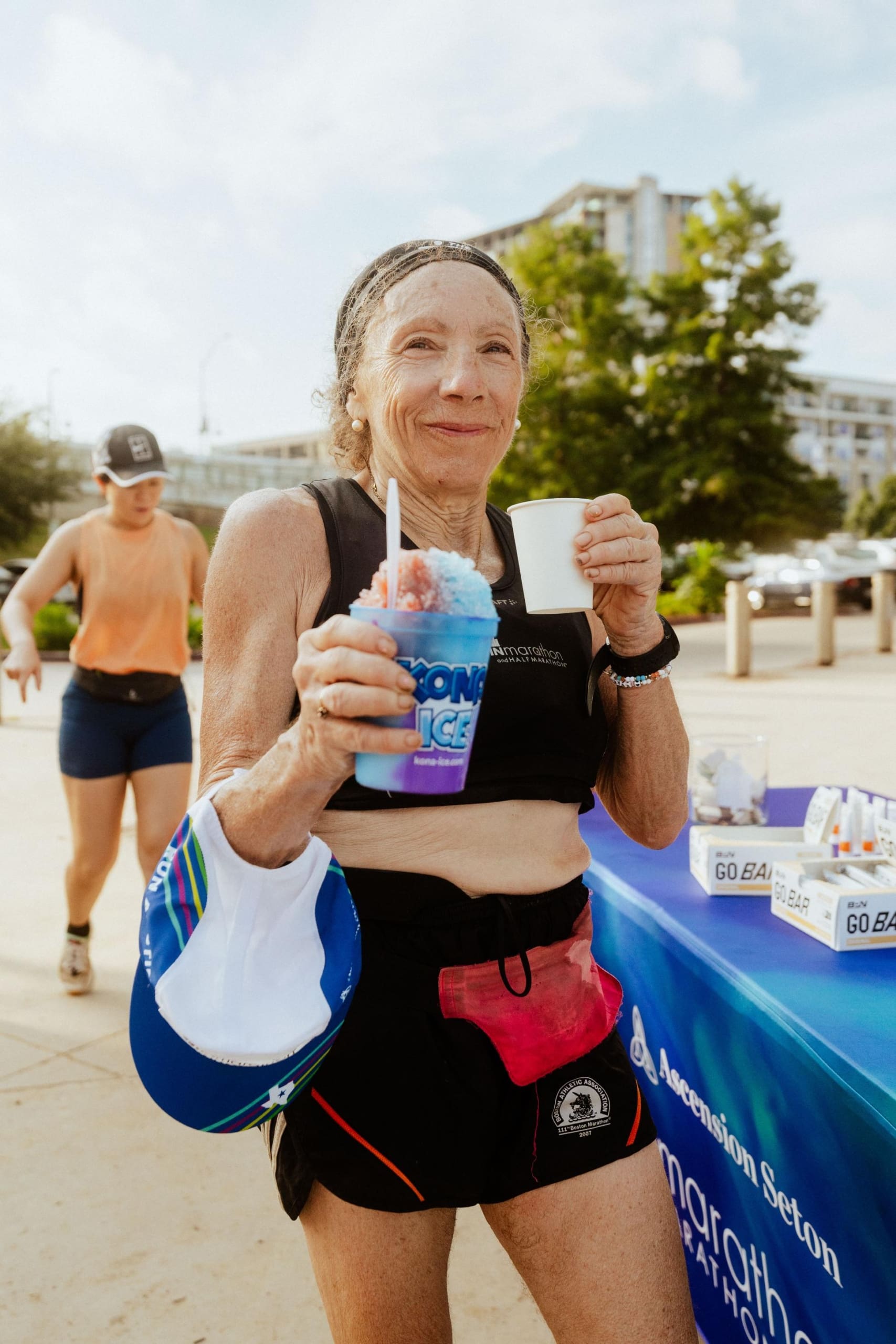 An older woman in athletic gear smiles while holding a snow cone and a cup, standing outdoors near a table with snacks and drinks. Another person in athletic wear walks behind her—enjoying Snow Cones on Trail on a sunny day. Austin Marathon Half Marathon & 5K