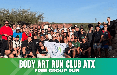 A large, diverse group of people pose together outdoors, smiling and holding a "BA" flag. A green banner below reads: "Body Art Run Club ATX Free Group Run." They're celebrating Launch Week ahead of the Austin International Half. Trees and rooftops are visible behind them. Austin Marathon Half Marathon & 5K