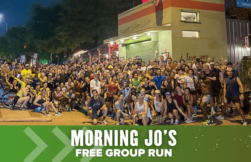 A large group of people pose together outside a building at night, many smiling and wearing running clothes. Text reads: "Morning Jo's Free Group Run" on a green banner, highlighting the excitement of this fun free run event. Austin Marathon Half Marathon & 5K