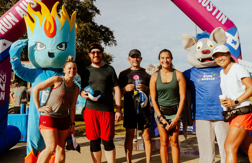 A group of five smiling runners pose under a purple archway with two mascots—one blue with flames and another dressed as a wolf—at an outdoor event, holding drinks and enjoying FREE Snow Cones while wearing athletic gear. Austin Marathon Half Marathon & 5K
