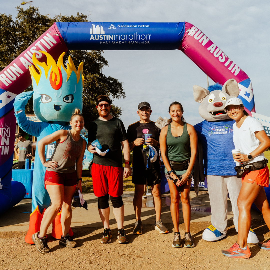 Six smiling runners pose under a colorful "Run Austin" arch, flanked by two mascots—one blue with a flame head and another gray with large ears. Everyone looks happy, dressed in athletic gear, ready to enjoy Snow Cones on Trail after the race. Austin Marathon Half Marathon & 5K