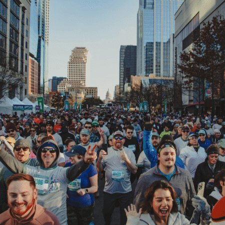 A large crowd of runners fills a city street during the 2026 Austin Marathon, surrounded by tall buildings. Many people are smiling, cheering, and wearing race bibs and sunglasses on a clear day. Austin Marathon Half Marathon & 5K