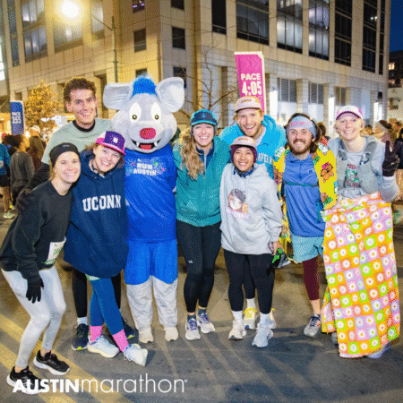 A group of smiling marathon runners pose together, some in colorful outfits and one in a blue mouse mascot costume, on a city street during the Austin Marathon in May. The atmosphere is cheerful and energetic, capturing the spirit of marathon training. Austin Marathon Half Marathon & 5K