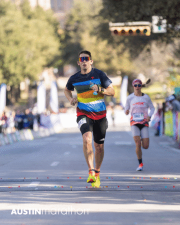 A man in a colorful striped shirt and sunglasses runs toward the finish line at the Austin Marathon race, with another runner behind him and blurred spectators in the background—captured in memorable Official Race Photos. Austin Marathon Half Marathon & 5K