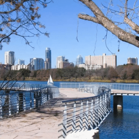 Curved boardwalk over a lake with metal railings, a small sailboat on the water, tree branches in the foreground, and a city skyline with tall buildings—perfect scenery for a relaxing day trip on a clear day. Austin Marathon Half Marathon & 5K