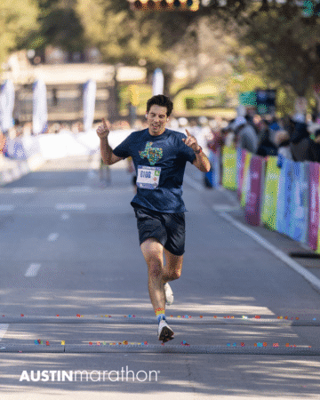 A male runner in a navy shirt and shorts crosses the finish line at the Austin Marathon race, arms raised in celebration, with confetti on the ground and spectators lining colorful barriers, ready to capture personal official race photos. Austin Marathon Half Marathon & 5K