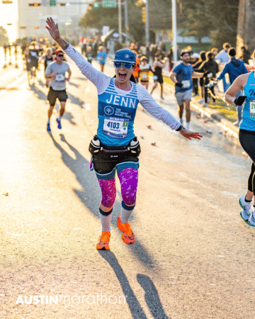A smiling runner in colorful gear with "JENN" on her shirt spreads her arms wide during the Austin Marathon race, captured in vibrant Official Race Photos with other runners and spectators cheering along the sunlit street. Austin Marathon Half Marathon & 5K