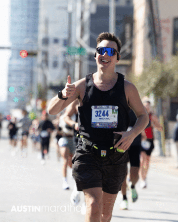 A smiling runner wearing sunglasses, a bib labeled "3244 Michael," and a black tank top gives a thumbs-up during the 2023 Austin Marathon race. Other runners and city buildings appear in the background, creating a lively scene perfect for Official Race Photos. Austin Marathon Half Marathon & 5K