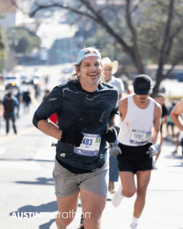 A smiling man in a dark shirt and cap runs in the Austin Marathon race, wearing bib number 1483. Other runners and a sunny, tree-lined street are visible in the background. This moment captures an inspiring personal achievement. Austin Marathon Half Marathon & 5K