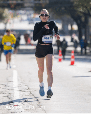 A runner wearing sunglasses, a black long-sleeve shirt, and shorts, with bib number 3764, competes in the Austin Marathon race. Other runners and orange traffic cones are visible in this Official Race Photo background. Austin Marathon Half Marathon & 5K